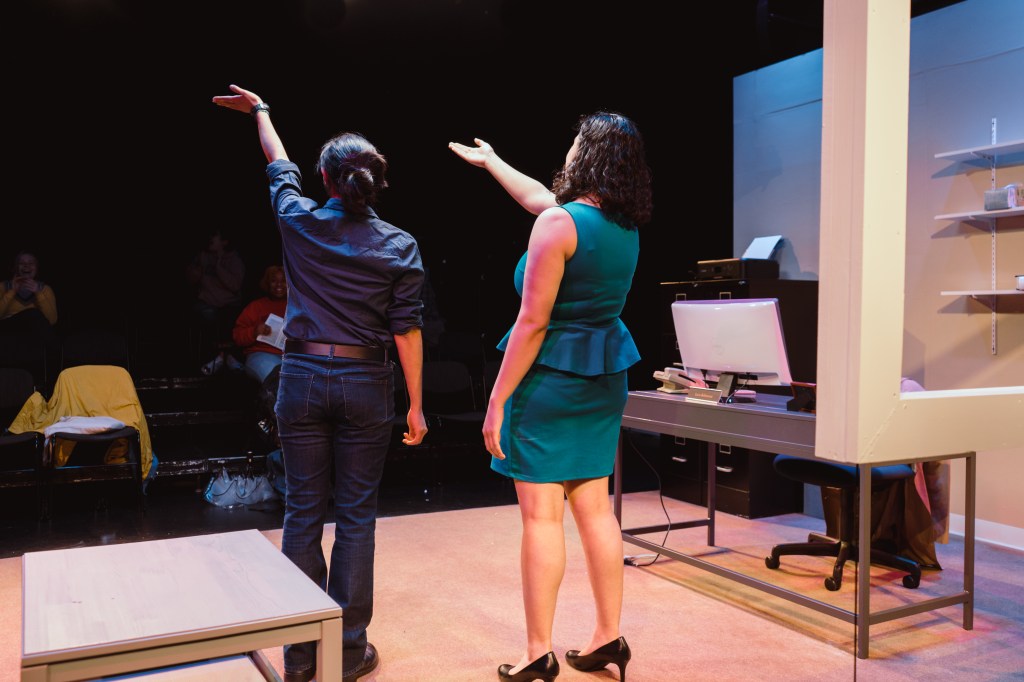 Image of two people on stage holding their hands out toward the audience