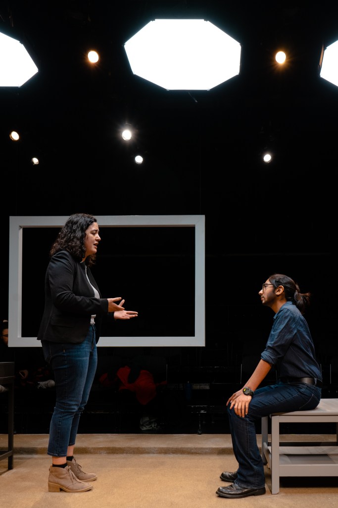 Image of two actors on stage with a square prop behind them
