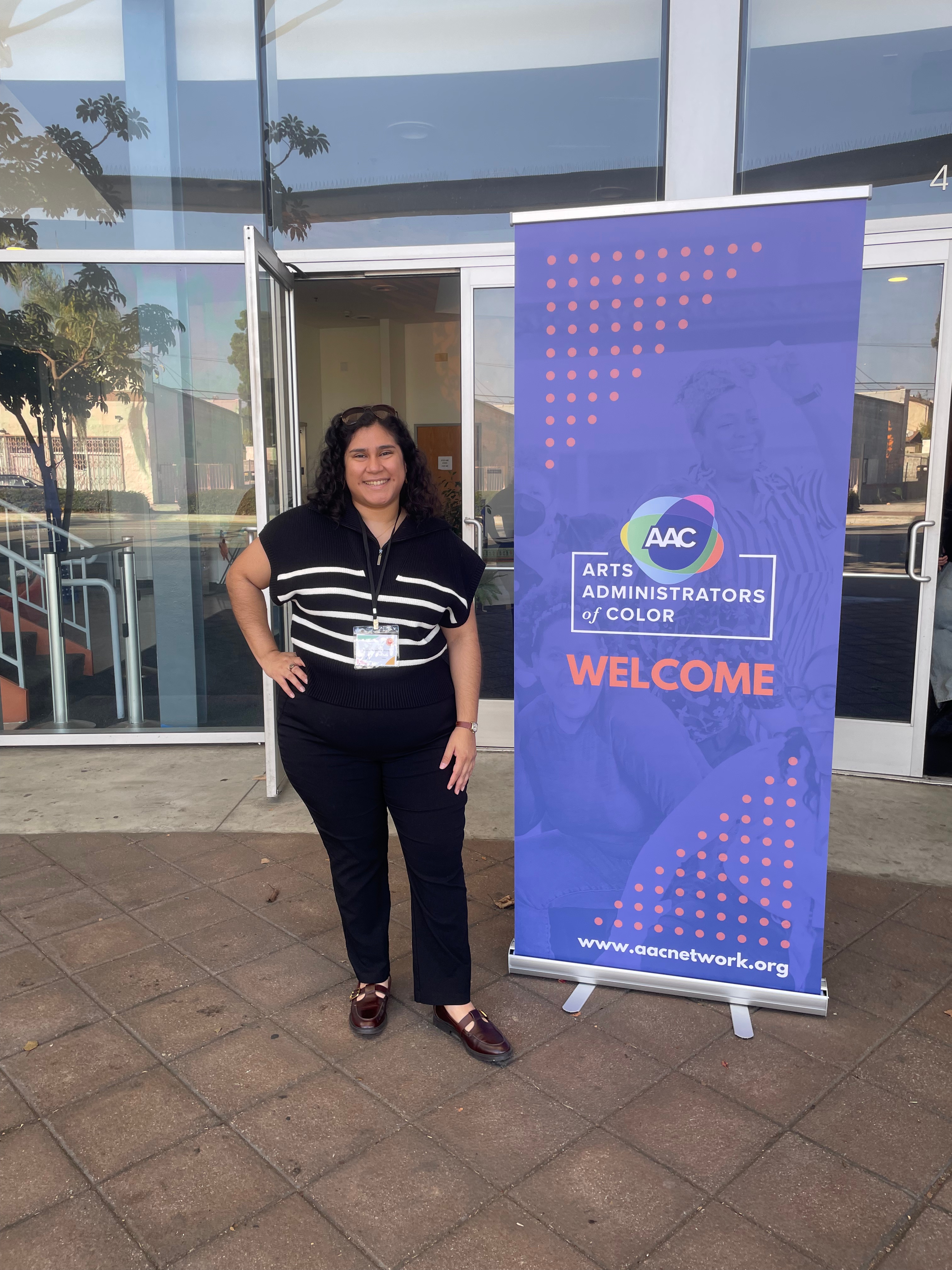 Alicia standing outside of a building next to an "Arts Administrators of Color" banner. Alicia is wearing a black top with white strips, black slacks, and brown shoes. The banner has the Arts Administrators of Color and is blue with a bit of orange