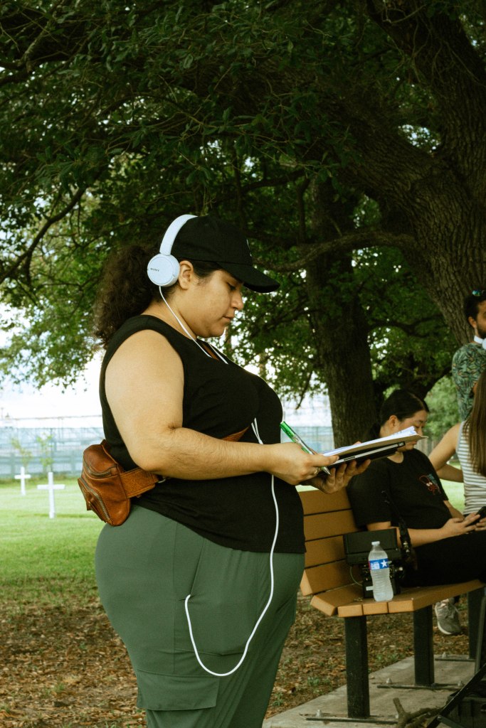 Alicia is outside, looking down at their notes and is using white headphones. Alicia is wearing a black tank top, a brown fanny pack, and green pants. Photo credit: @yenni.coolpix