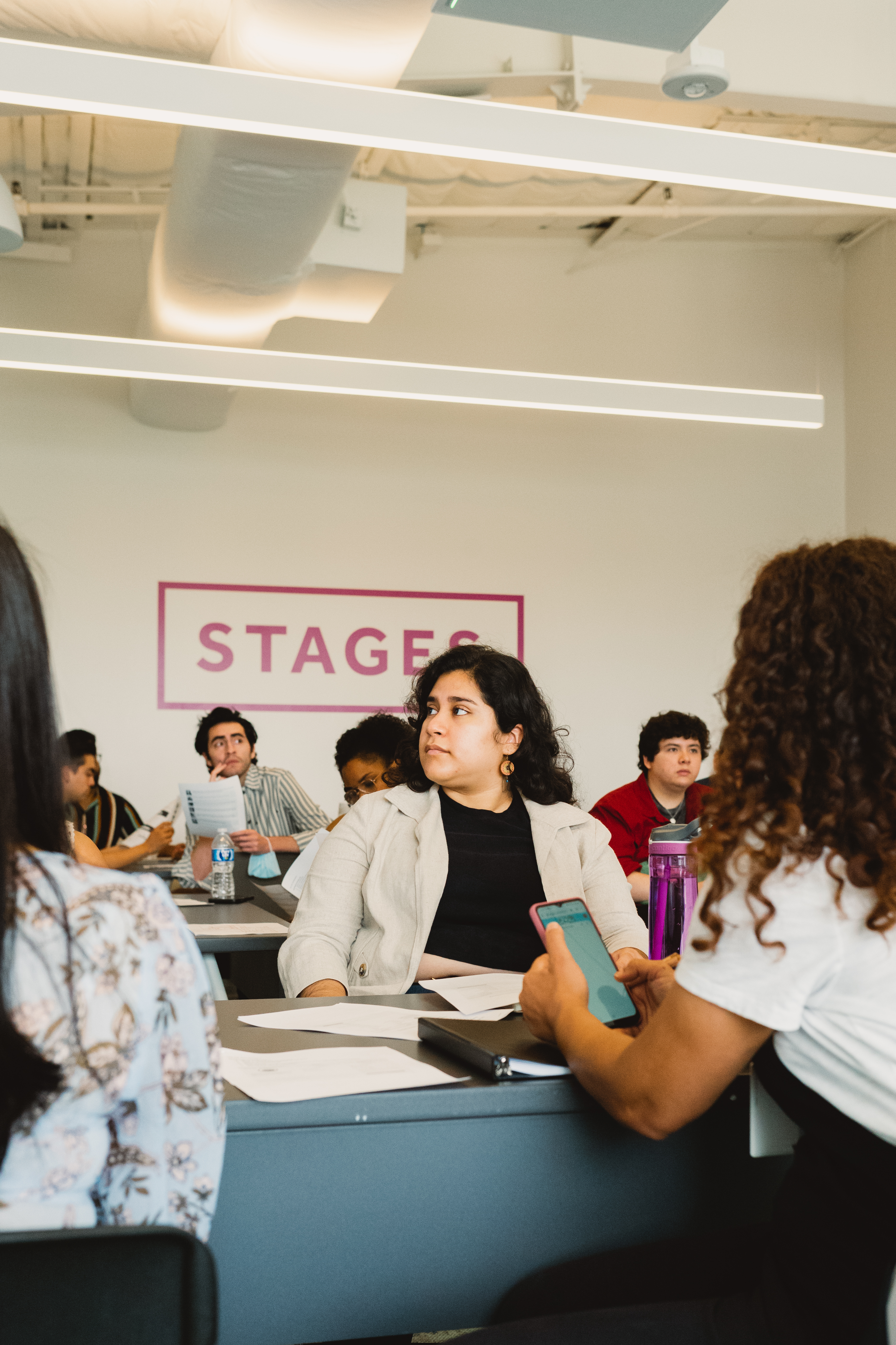 Picture of Alicia at a stages workshop. Alicia face is turned slightly to the left and is listening to the speaker. There are about 6 people in the background listening to the speaker, working and interacting with each other. Photo credit: gvelasquez (2023)