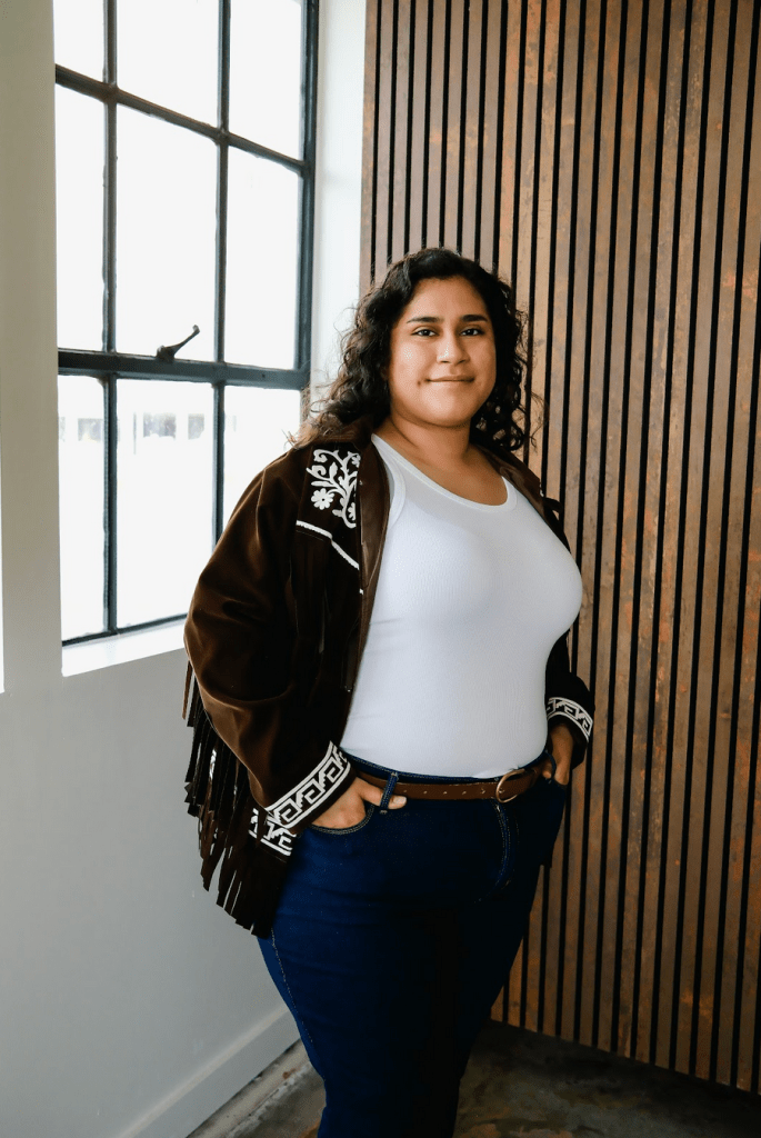 Head shot of Alicia, who is looking at the camera with a slight smile. Alicia is wearing a dark brown jacket with fringe on the side, a white tank top, brown belt, and dark colored jeans. Photo credit: Sarah Ontiveros