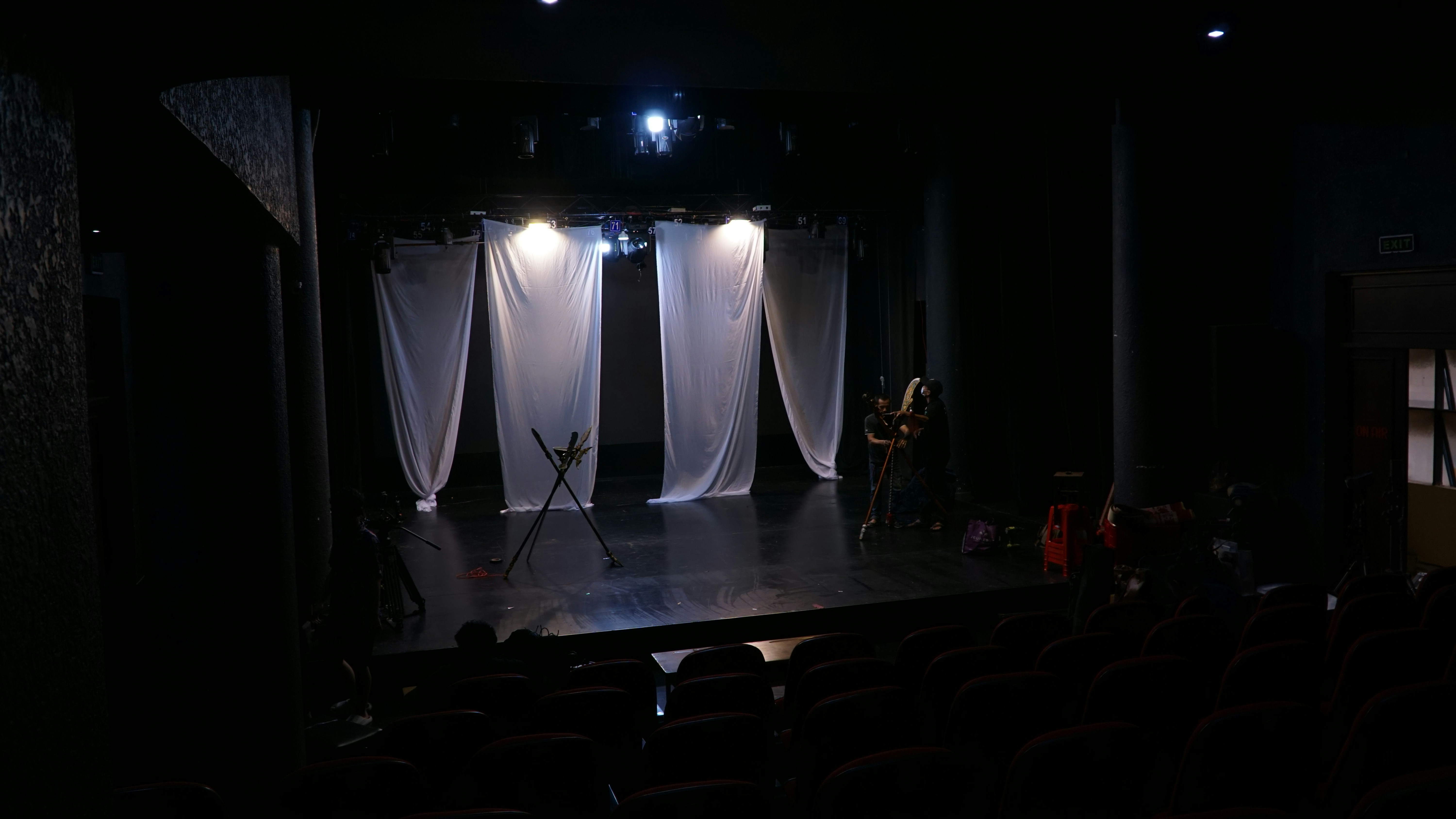 Image of a mostly-empty stage with white curtains hanging down and two crew members 